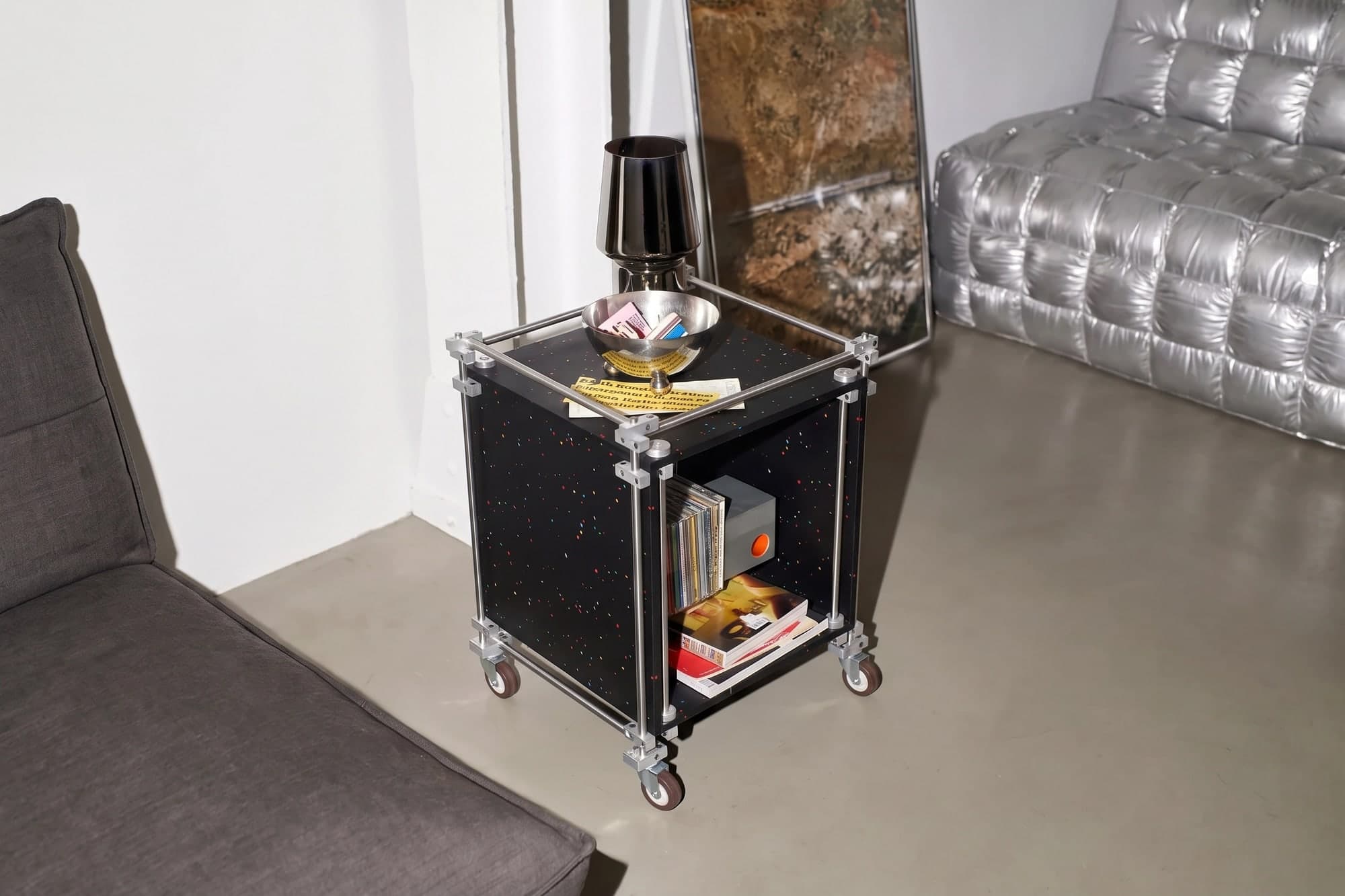 A cube trolley in black recycled polystyrene with colour flecks, on steel rods, in a room